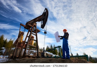 Full Length Of Oil Man In Work Overalls And Helmet Holding Sheet Of Paper While Standing Near Oil Pumping Unit. Male Worker Studying Plan Of Oil Field, Checking Work Of Pump Jack Under Beautiful Sky.