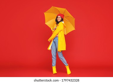 Full length optimistic ethnic woman with umbrella smiling and looking at camera on rainy autumn day against red background