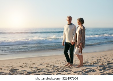 Full length outdoor shot of loving senior couple walking along the beach in evening. Mature man and woman strolling during sunset on the sea shore.