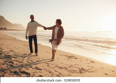 Full length outdoor shot of playful senior couple walking along the beach at sunset. Mature man and woman walking on the sea shore.