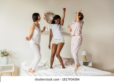 Full Length Overjoyed African American Young Woman Having Fun With Crazy Asian Korean And European Appearance Female Best Friends, Jumping Dancing To Favorite Music On Bed, Feeling Energetic On Party.