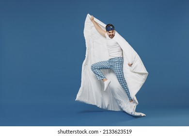 Full length overjoyed fun winner young man 20s in pajamas jam sleep mask resting relaxing at home jump high flying with duvet blanket isolated on dark blue background. Good mood night bedtime concept.