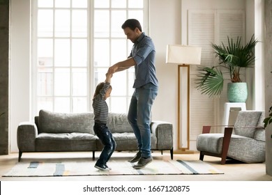 Full Length Overjoyed Little Preschool Kid Boy Holding Hands Of Smiling Young Father, Jumping Together In Living Room. Happy Dad Dancing To Music, Having Fun With Energetic Small Child Son At Home.