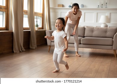 Full Length Overjoyed Little Preschool Cute Vietnamese Ethnic Baby Girl Running Barefoot On Warm Floor, Having Fun Playing With Energetic Young Asian Mother Or Nanny, Leisure Daycare Activity.