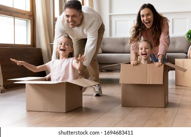 Full length overjoyed little sisters sitting in big cardboard boxes, playing with happy parents in new home. Excited laughing family couple having fun together with adorable daughters in living room.