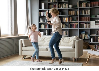 Full length overjoyed middle aged senior grandmother in eyeglasses dancing to favorite disco music with energetic little preschool cue granddaughter in living room, family domestic activity concept.