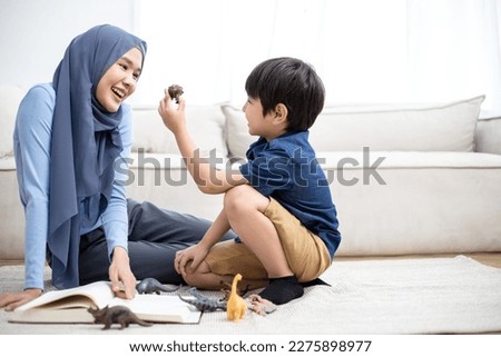 Full length overjoyed muslim family of two playing with dinosaurs toys on table. Happy young mommy babysitter having fun with cute preschool kid boy in living room