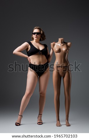 full length of overweight young woman in swimsuit and sunglasses posing with plastic mannequin on grey