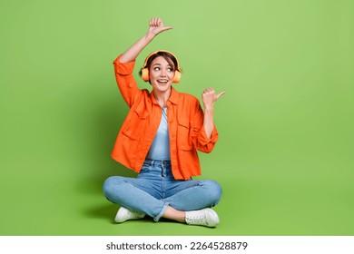 Full length photo of adorable excited woman wear denim jacket headphones thumbs empty space isolated green color background