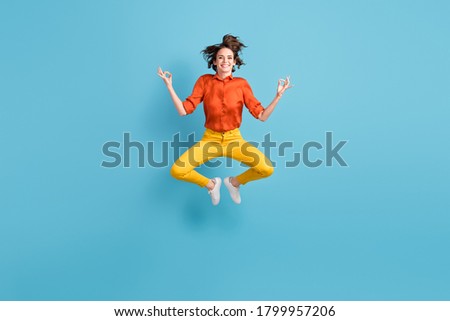 Full length photo of attractive funny lady jump high up positive energy emotions practice yoga om position in flight wear orange shirt yellow trousers sneakers isolated blue color background