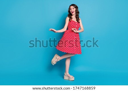 Full length photo of attractive pretty curly lady good mood flirty raise leg hands dancing look side wear summer red white dotted retro dress open toes shoes isolated blue color background