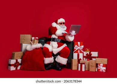 Full length photo of bossy santa sit buy gifts online wear cap glasses coat pants footwear isolated on red background - Powered by Shutterstock