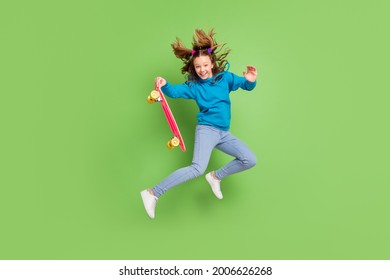 Full length photo of brunette ponytail haired girl jump wear jeans hold longboard fly hair isolated on green color background