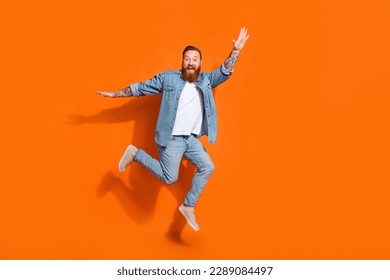 Full length photo of cheerful funky guy dressed denim jacket jumping high empty space isolated orange color background