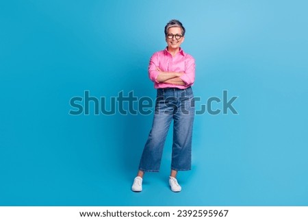 Full length photo of cheerful optimistic woman dressed pink shirt denim trousers holding arms folded isolated on blue color background