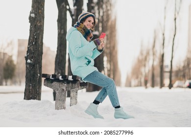 Full length photo of cheerful positive young woman look phone message hold coffee sit bench in park outdoors