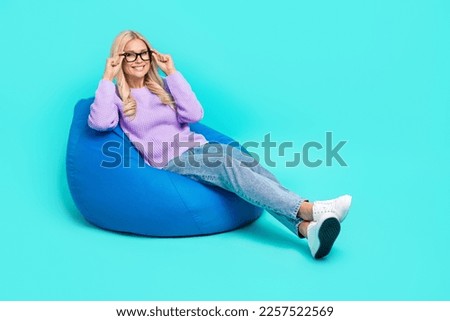 Full length photo of cheerful pretty lady sit cozy bag hands touch eyeglasses isolated on cyan color background