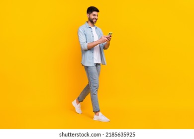 Full length photo of cheery hispanic man walking hold telephone chatting dressed stylish denim look isolated on yellow color background
