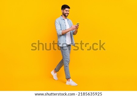 Full length photo of cheery hispanic man walking hold telephone chatting dressed stylish denim look isolated on yellow color background