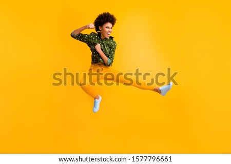Full length photo of crazy dark skin curly lady jumping high making fighting moves ready to kick leg and fist wear green dotted shirt trousers isolated yellow color background