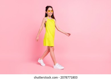 Full length photo of cute charming school girl wear yellow dress dark eyewear walking empty space isolated pink color background