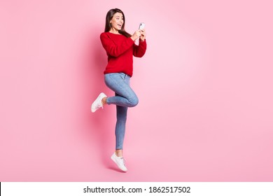 Full length photo of cute girl jump excited screen hold telephone wear red sweater denim jeans sneakers isolated pink color background