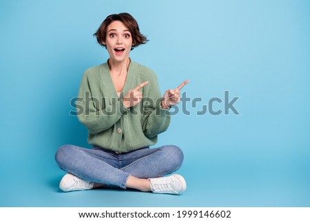 Full length photo of cute impressed young girl dressed green cardigan sitting floor pointing empty space isolated blue color background