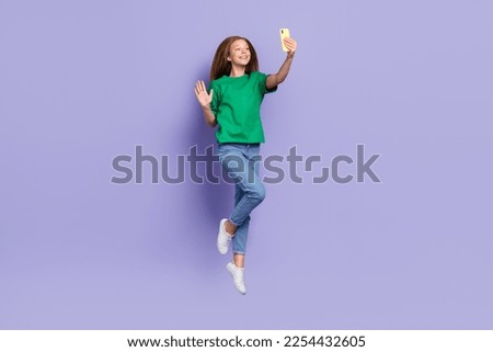 Full length photo of cute sweet girl dressed green t-shirt jumping high waving arm palm modern device isolated purple color background