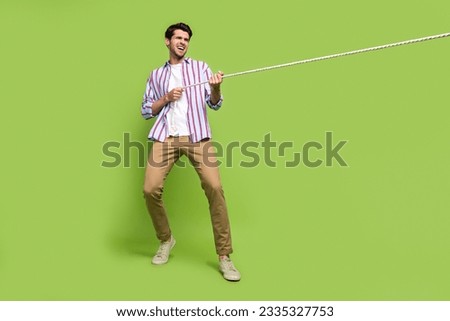 Full length photo of determined man with stubble dressed striped shirt pull rope from empty space isolated on green color background
