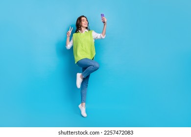 Full length photo of excited carefree girl jumping make selfie show v-sign isolated on blue color background