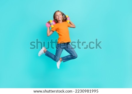 Full length photo of excited cool little child dressed orange t-shirt jumping high rising skateboard isolated teal color background