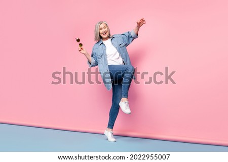 Full length photo of fancy senior lady go hold spectacles wear jeans shirt isolated on pink background