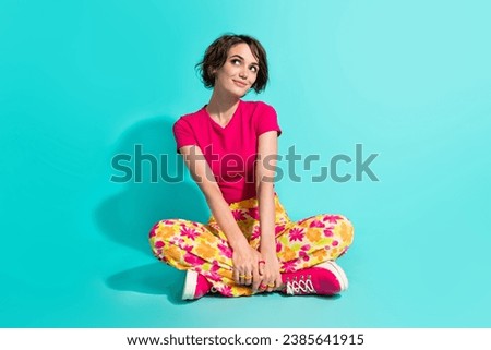 Full length photo of funky dreamy woman dressed flower print trousers sitting legs crossed looking empty space isolated teal color background