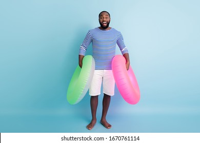 Full length photo of funny dark skin guy holding two green pink life buoy ready to swim ocean sea wear striped sailor shirt white shorts isolated blue color background