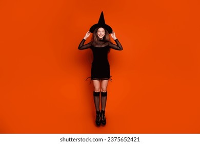 Full length photo of funny excited girl dressed dark witch dress headwear jumping high isolated orange color background
