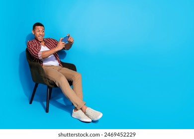 Full length photo of funny guy wear stylish t-shirt sit on armchair look at smartphone play video game isolated on blue color background - Powered by Shutterstock