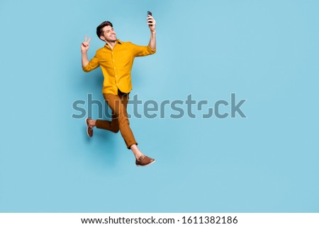Full length photo of funny guy jumping high holding telephone making selfies showing v-sign symbol wear yellow shirt pants isolated blue color background