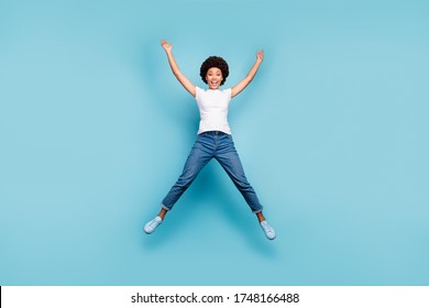 Full length photo of funny rejoicing dark skin wavy lady jumping high sunny summer day cheerful good mood wear casual white t-shirt jeans shoes isolated blue color background - Shutterstock ID 1748166488
