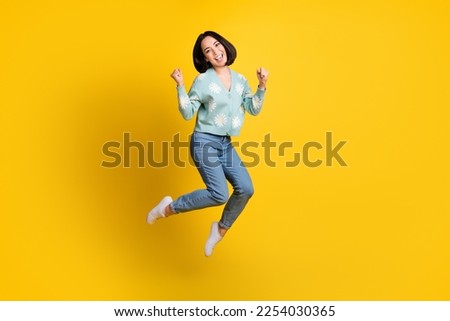 Full length photo of girlish nice satisfied girl wear blue cardigan jeans clenching fists jumping isolated on yellow color background