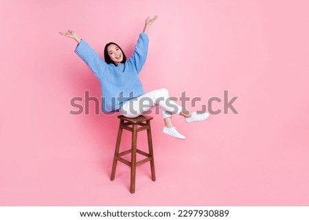 Full length photo of gorgeous pretty girl blue sweater white pants sitting on bar stool raising arms up isolated on pink color background