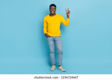 Full Length Photo Of Handsome Dude Showing V-sign Hippie Symbol Wear Yellow Pullover Jeans Sneakers Isolated Blue Color Background