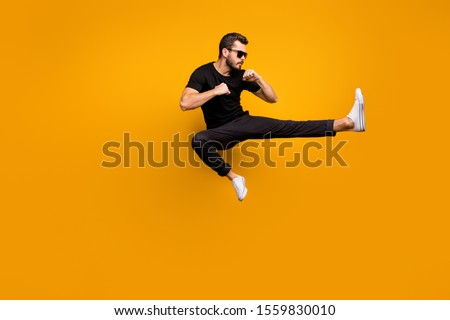 Full length photo of handsome guy jumping high practicing self defense kicking confident facial expression wear sun specs black t-shirt pants isolated yellow color background