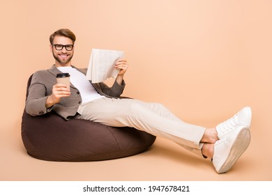 Full length photo of happy charming young man sit chair hold newspaper cup isolated on beige color background - Powered by Shutterstock