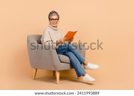Full length photo of happy old grandmother spending her pensioner like dream with book sitting armchair isolated on beige color background