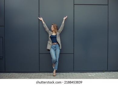 Full length photo of happy young businesswoman standing on one leg leaning on gray wall of office building with raised arms. Urban lifestyle, entrepreneur, well dressed lady, successful people.