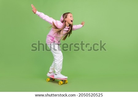 Full length photo of impressed little blond girl ride skate wear hoodie trousers sneakers isolated on green background