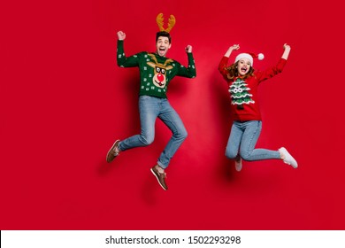 Full length photo of lady and guy jumping excited by x-mas discounts wear ugly ornament jumpers and headwear isolated red color background