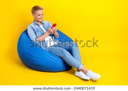Full length photo of lovely young lady sit bean bag hold device write comment wear trendy jeans garment isolated on yellow color background