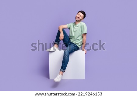 Full length photo of nice young man sitting white copyspace cube cheerful dressed stylish gray clothes isolated on purple color background