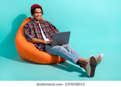 Full length photo of optimistic clever guy wear plaid shirt beanie hat sit on bean bag with laptop isolated on turquoise color background - Powered by Shutterstock